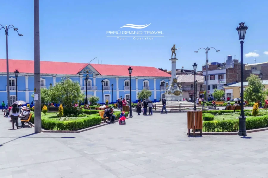 Parque Pino em Puno, no Peru, com jardins, bancos e o monumento central, cercada por edifícios coloniais sob um céu andino azul e limpo.