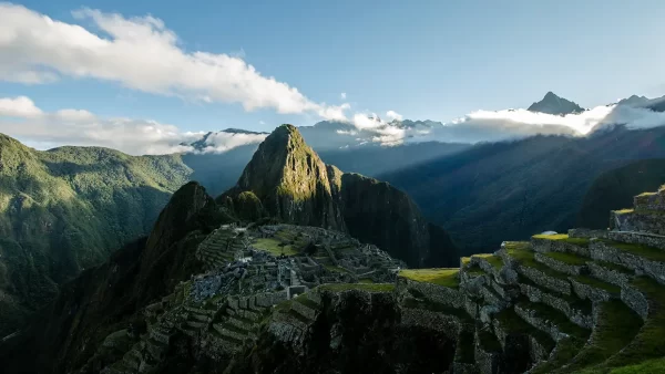 Machu Picchu com céu azul e o sol nascendo, vista dos terraços, das construções incas e na parte inferior à esquerda o rio Urubamba e muitas montanhas que cercam Machu Picchu