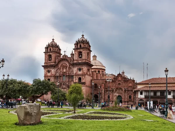 A cidade de Cusco na Plaza de Armas com vista para a Igreja da Companhia de Jesus, diferentes pessoas na praça, caminhando ou sentadas, várias árvores e um jardim verde.