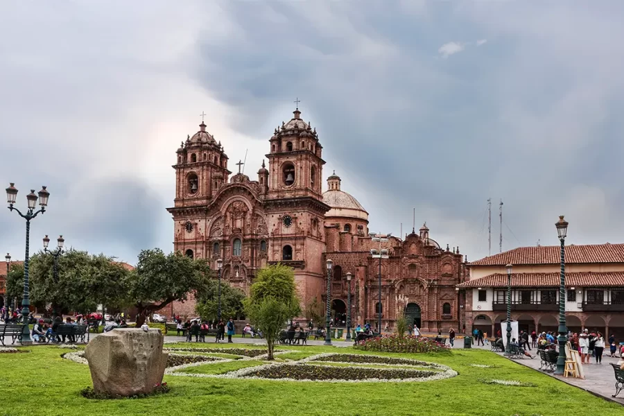 A cidade de Cusco na Plaza de Armas com vista para a Igreja da Companhia de Jesus, diferentes pessoas na praça, caminhando ou sentadas, várias árvores e um jardim verde.