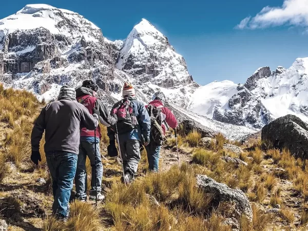 Turistas caminhando em direção a uma montanha nevada, com seus bastões de caminhada e mochilas ao longo de um caminho de paralelepípedos e vistas de paisagens nevadas
